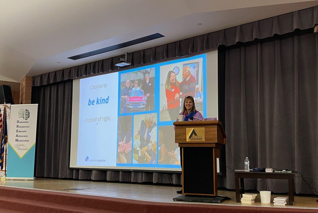 Executive Director of Dementia Together, Cyndy Luzinski, stands behind a podium on a stage at Alpena Community College.